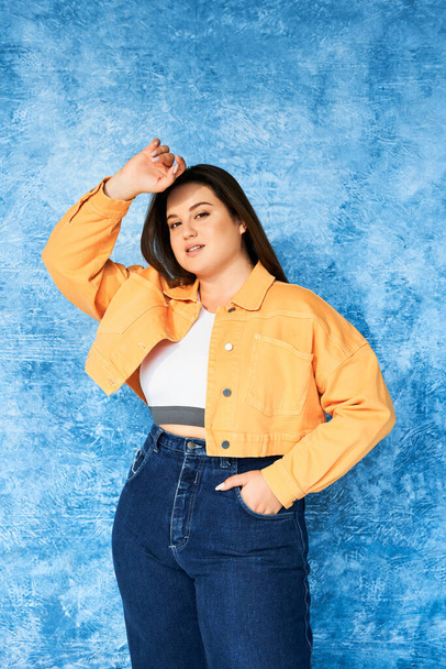 plus size woman with long hair and natural makeup wearing crop top, orange jacket and denim jeans while posing with hand in pocket and looking at camera on mottled blue background, body positive  - Fotó, kép