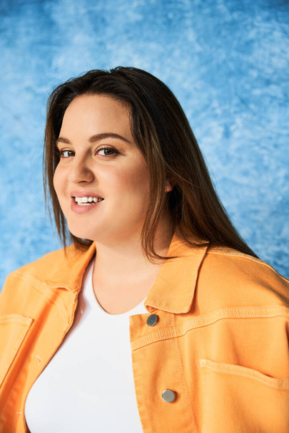 portrait of positive plus size woman with long hair and natural makeup wearing crop top and orange jacket while posing and looking at camera on mottled blue background, body positive  - Fotó, kép