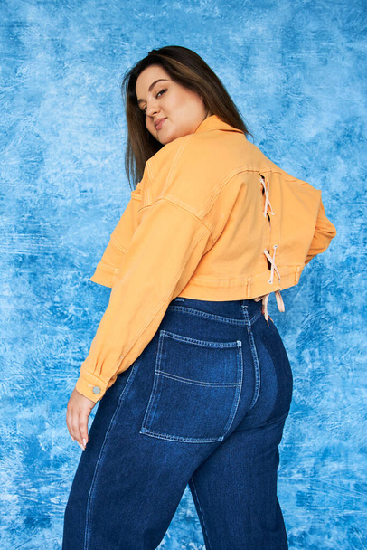 alluring plus size woman with long hair and natural makeup wearing crop top, orange jacket and denim jeans while posing and looking at camera on mottled blue background, body positive  - Foto, imagen