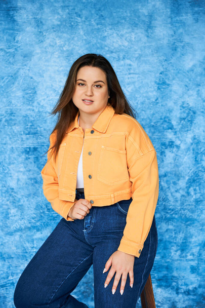 barna plusz méret nő hosszú haj és természetes smink visel narancs dzseki és farmer, miközben ül a széken, és nézi a kamera foltos kék háttér, test pozitív  - Fotó, kép