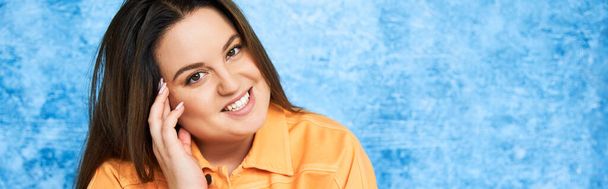 smiling plus size woman with brunette hair and natural makeup wearing orange jacket and touching cheek while posing and looking at camera on mottled blue background, body positive, banner  - Photo, Image