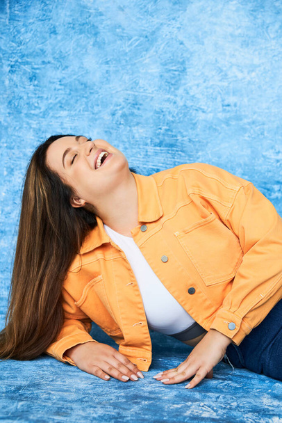 body positive and happy plus size woman with long hair and natural makeup laughing with closed eyes while posing in orange jacket and denim jeans on mottled blue background  - Photo, Image
