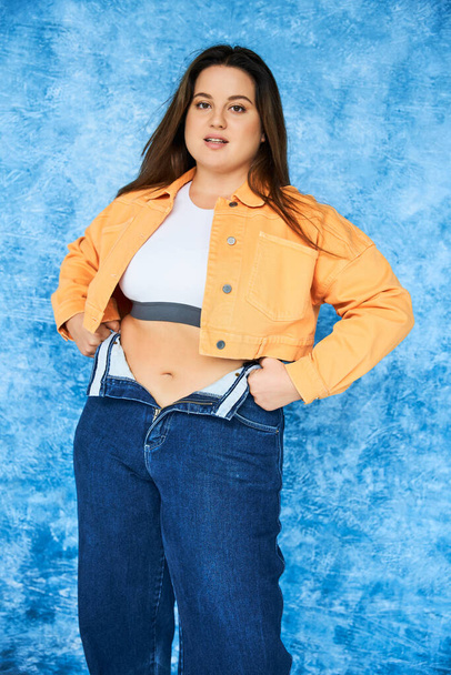 body positive and brunette plus size woman with long hair and natural makeup, in crop top and orange jacket wearing denim jeans while posing and looking at camera on mottled blue background  - Fotó, kép