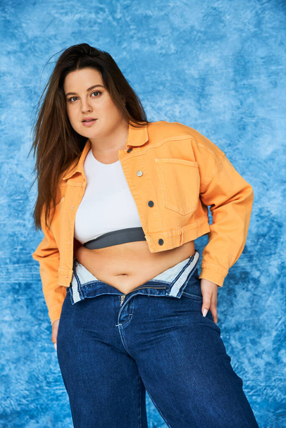 body positive and brunette plus size woman with long hair and natural makeup posing in crop top, orange jacket and denim jeans while standing and looking at camera on mottled blue background  - Fotó, kép