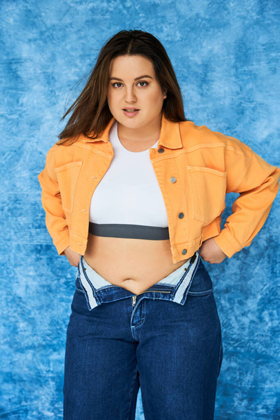 body positive woman with long hair and natural makeup posing in crop top and orange jacket while wearing denim jeans while standing and looking at camera on mottled blue background  - Photo, Image
