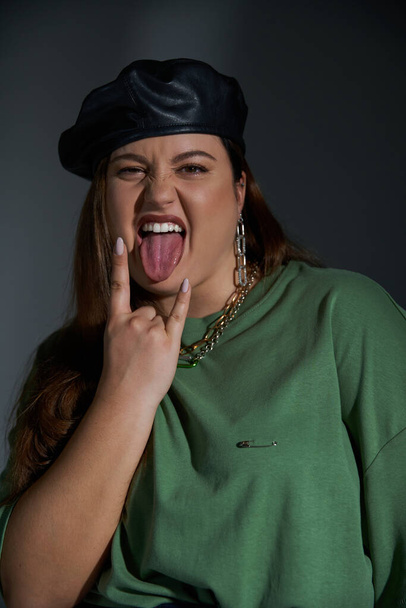 portrait of stylish plus size woman posing in leather beret and green t-shirt, looking at camera while sticking out tongue and showing rock sign on dark background with studio lighting  - Фото, изображение