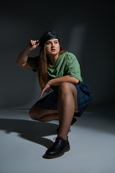 Frau in voller Länge posiert in Lederbarett, grünem T-Shirt, kariertem Rock mit Ketten, Netzstrumpfhose und schwarzen Schuhen sitzend und wegschauend auf dunklem Hintergrund mit Studiobeleuchtung  - Foto, Bild