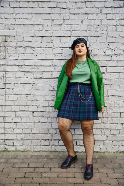 full length of chic plus size woman posing in plaid skirt with chains, green leather jacket, beret, fishnet tights and black shoes while posing near brick wall on urban street, body positive  - Photo, Image