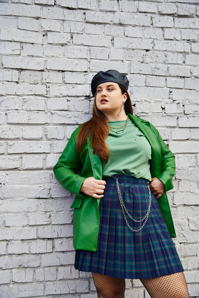 chic plus size vrouw poseren in geruite rok met kettingen, groene leren jas, zwarte baret en visnet panty terwijl weg te kijken en staan in de buurt van baksteen muur op stedelijke straat, lichaam positief  - Foto, afbeelding