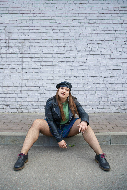 femme à la mode plus la taille posant en veste et béret en cuir, jupe à carreaux, collants résille et chaussures noires tout en posant près du mur de briques sur la rue urbaine, corps positif, pleine longueur  - Photo, image
