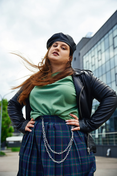 confident woman with plus size body posing in leather jacket with black beret near blurred modern building on urban street outdoors, body positive, hands on hips, looking at camera - Photo, Image