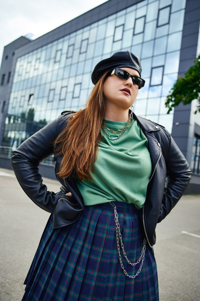 brunette woman with plus size body posing with hands on hips while standing in stylish sunglasses, leather jacket with black beret, plaid skirt near blurred modern building on urban street  - Photo, Image
