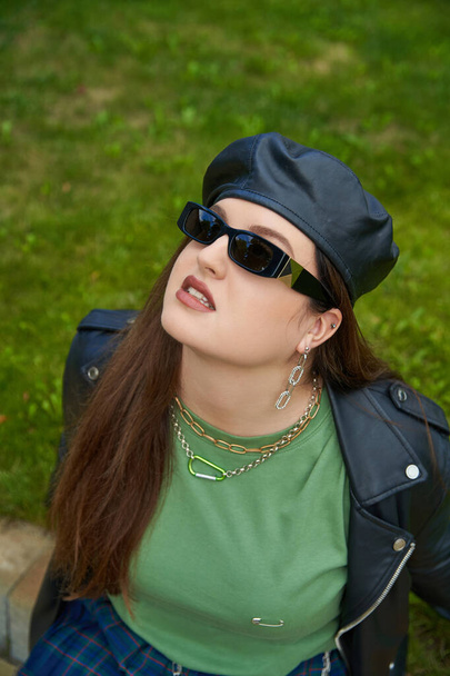 high angle view of plus size woman in sunglasses, leather jacket with black beret and chain accessories looking up in green park with fresh lawn, body positive, outdoors  - 写真・画像