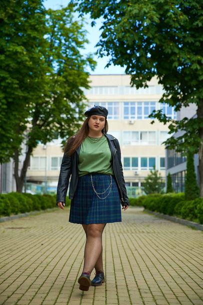 plus size woman walking in leather jacket, beret, plaid skirt, fishnet tights and black shoes while looking at camera on urban street with building and trees on blurred background, full length  - Photo, Image