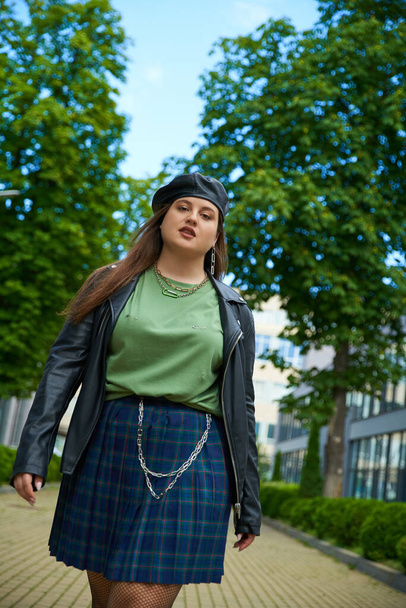 confident plus size woman walking in leather jacket with black beret, plaid skirt and green t-shirt near blurred modern building on urban street outdoors, body positive, trees on background  - Photo, Image