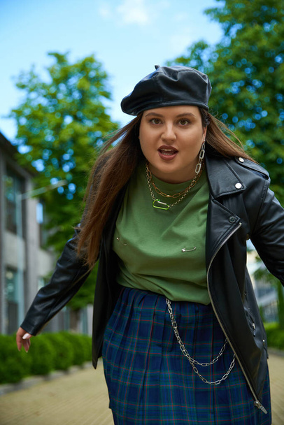 confident woman with plus size body looking at camera and posing in leather jacket with black beret near blurred modern building and green trees on urban street outdoors, body positive  - Photo, Image