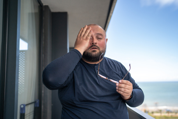 Suffering overweight man feeling headache rubbing eyes stands on balcony on hot weather in summer. Sweating tired guy suffer pressure problems living in tropical climate high humidity. Health problems - Photo, Image