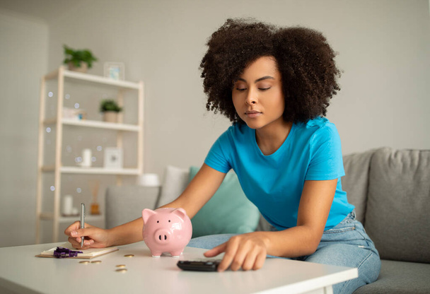 Concentrated millennial curly black lady counting income, tax and mortgage at table with piggy bank and calculator in living room interior. Debts, finance, home accounting - Foto, imagen
