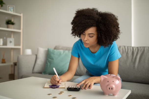 Serious young black curly lady makes accounting, make notes at table with piggy bank and calculator, counts profit in living room interior. Finances, paying taxes, savings at home - Foto, imagen