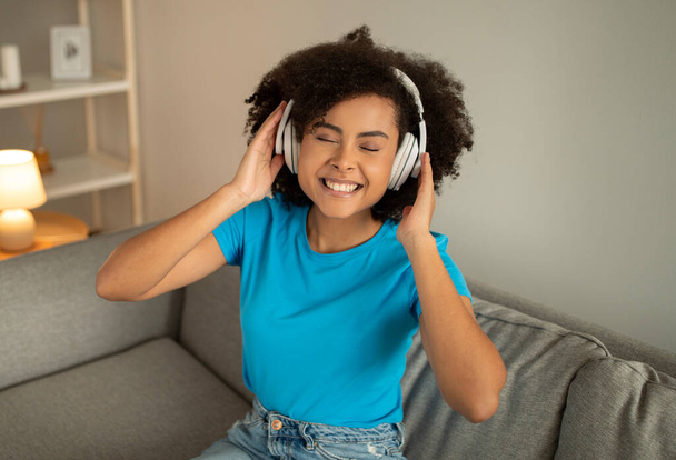 New gadget and audio app. Positive young black curly woman in wireless headphones sits on sofa, enjoys free time alone, dancing and listen music in living room interior. Break, rest at home - Photo, Image