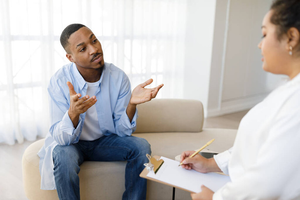Handsome millennial black guy in casual sitting on couch at therapist office clinic, sharing feelings and gesturing, young anxious man attend therapy session with woman psychologist, copy space - Fotó, kép
