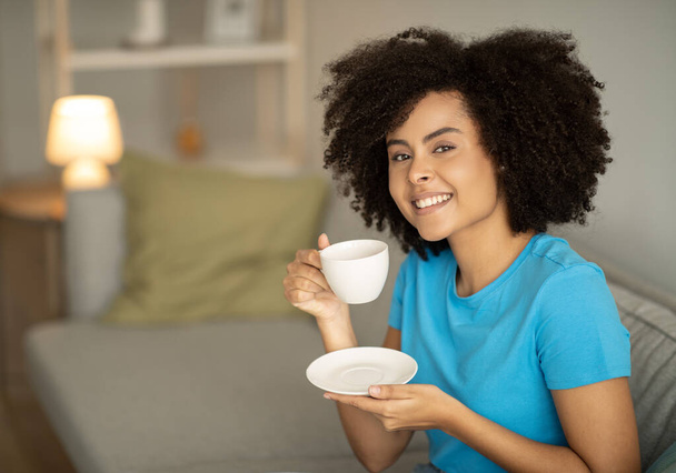 Good morning and fresh drink. Cheerful young black curly woman drinks cup of hot coffee on sofa, enjoy free time alone in evening, relax in living room interior. Tea and rest at home, ad and offer - 写真・画像