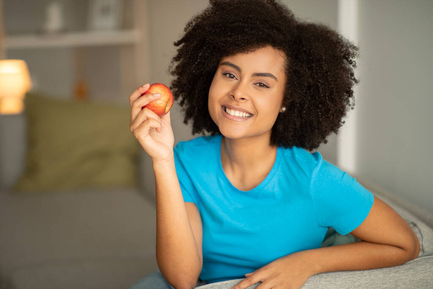 Cheerful millennial black curly woman holding apple on sofa, enjoy free time, perfect teeth in living room interior. Snack, vegetables diet, vitamins and proper nutrition at home, ad and offer - Foto, immagini
