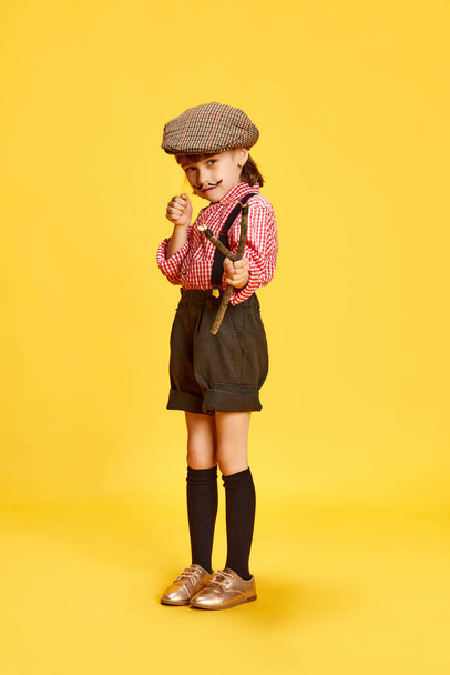 Portrait of little girl, child in retro clothes, shirt and shorts, holding slingshot against yellow studio background. Concept of childhood, emotions, fun, fashion, lifestyle. Outdoor game - Photo, Image