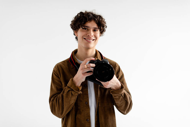 Επαγγελματίας φωτογράφος. Ευτυχισμένος όμορφος νεαρός με κομψό casual ντύσιμο χρησιμοποιώντας dslr κάμερα, που εργάζονται σε φωτογραφικό στούντιο, στέκεται πάνω από λευκό φόντο, αντιγραφή χώρου - Φωτογραφία, εικόνα