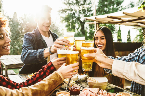 Zróżnicowani młodzi ludzie siedzący przy stole przy barze tosty szklanki piwa w browarnym ogrodzie pubowym. Happy hour, przerwa obiadowa i koncepcja młodzieży - Zdjęcie, obraz