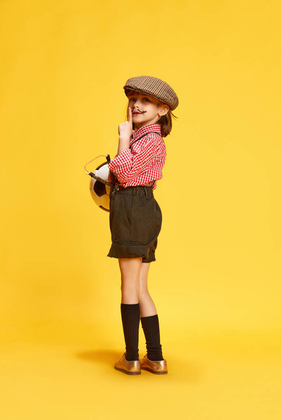 Portrait of little girl, child in shirt and shorts standing with football ball against yellow studio background. Keeping secrets. Concept of childhood, emotions, fun, fashion, lifestyle - Zdjęcie, obraz