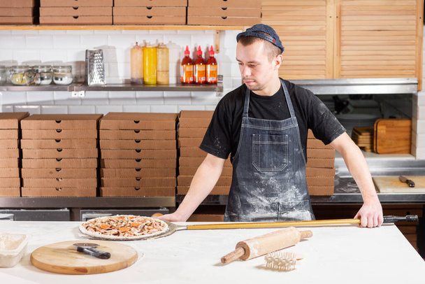 Primo piano mano dello chef panettiere in uniforme blu grembiule taglio pizza in cucina. Foto di alta qualità - Foto, immagini