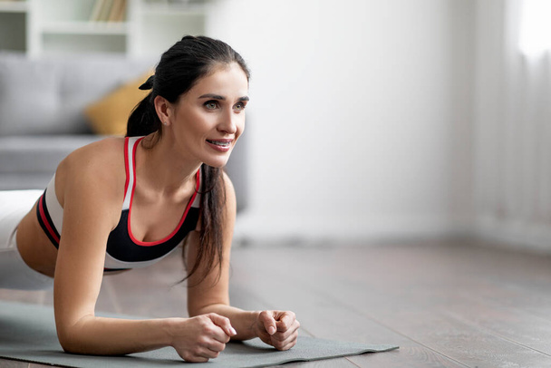 Closeup of motivated beautiful well-fit young brunette woman wearing sportswear standing on elbows on yoga mat, planking at home, looking at copy space. Workout at home, healthy fit body concept - Фото, зображення