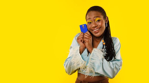 Εύκολη πληρωμή. Χαμογελώντας μαύρη κυρία κρατώντας πιστωτική κάρτα κοντά στην τράπεζα διαφήμισης πρόσωπο ή ψώνια προσφέρουν χαμογελώντας στην κάμερα ποζάροντας στο κίτρινο φόντο στούντιο. Πανόραμα με χώρο αντιγραφής - Φωτογραφία, εικόνα