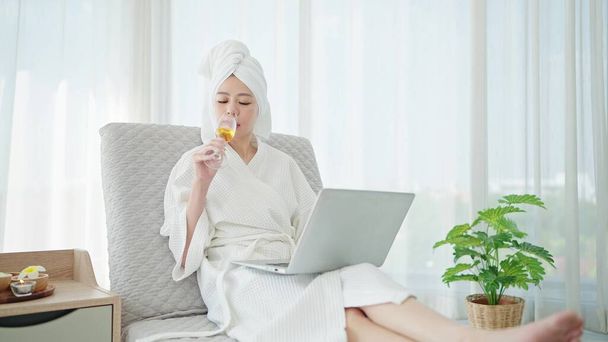 Ázsiai nő fürdőköpenyben és törölköző a fejét pezsgő bort iszik, miközben dolgozik a laptop és pihen a kanapén luxus hálószoba otthon - Fotó, kép