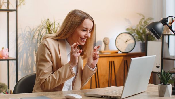 Młoda kobieta biznesu pracuje na laptopie w biurze domu biurko skrzyżowane palce patrząc na ekranie z prośbą o szczęście wiadomości życząc dobrych wyników egzaminu, marząc o zwycięstwie w loterii jackpot - Zdjęcie, obraz