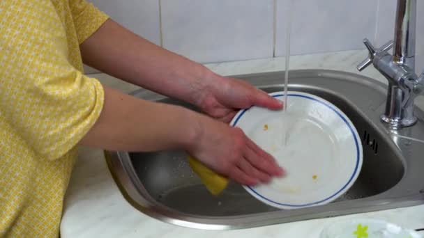 Vrouw handen met gele spons was vaatwerk in wastafel close-up. Huisvrouw maakt schoon van plaat onder stromend water. Schoonmaakster schoonmaken huishoudelijke klusjes keukengerei. Thuis afwassen. Huishouding. - Video