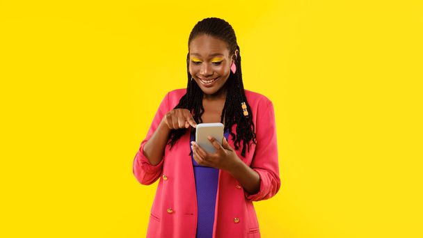 Happy Tech Savvy African American Woman Navegando na Internet no telefone, Envolvendo-se na diversão de mídia social com aplicativos de smartphones em pé sobre fundo amarelo, vestindo roupas brilhantes e maquiagem. Panorama - Foto, Imagem