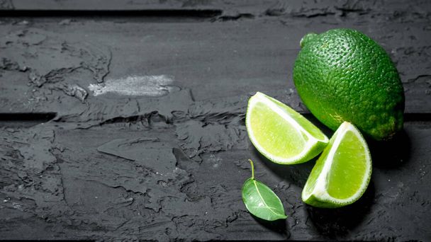 Pieces of fresh lime and one whole lime. On black rustic background - Zdjęcie, obraz