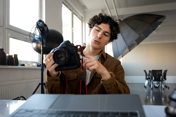 European male photographer using modern photo camera while working on laptop at workplace in studio. Professional photography and creative business career concept - Foto, imagen