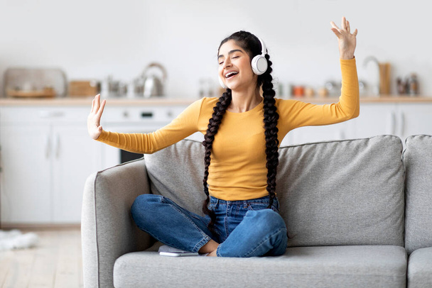 Plezier thuis. Portret van vrolijke jonge Indiase vrouw Luisteren haar favoriete muziek in draadloze hoofdtelefoon, zorgeloze Oosterse vrouw dansen met de handen terwijl zitten op de bank in de woonkamer - Foto, afbeelding
