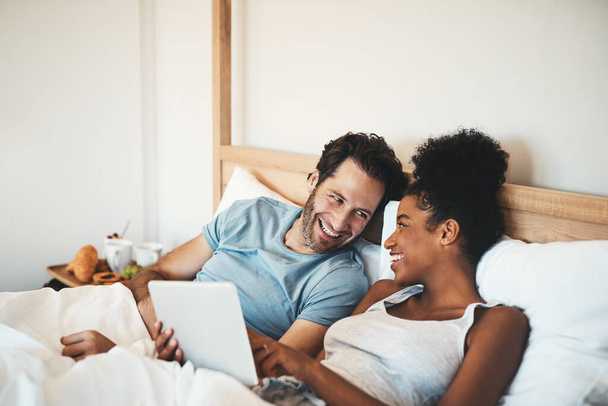 Happy couple, tablet and laughing on bed for joke, funny entertainment or online streaming at home. Interracial man and woman person on technology in relax for morning or social media in the bedroom. - Фото, зображення