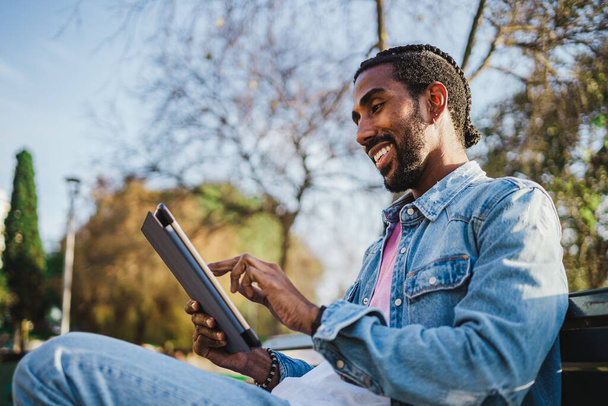 Lächelnder junger Mann beim Lesen eines Buches auf seinem digitalen Tablet. Er sitzt auf einer Bank im Park. - Foto, Bild