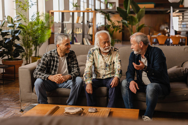 três velhos amigos conversando no sofá em casa, reunião de homens seniores com cabelos grisalhos se divertindo dentro de casa. - Foto, Imagem