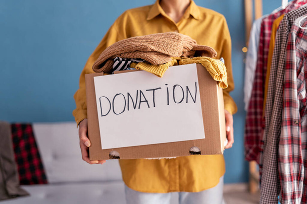 Spendenkonzept. Frau hält Spendenbox mit Spendenkleidung in ihrer Wohnung. Wohltätigkeit. Armen Menschen helfen. Nahaufnahme - Foto, Bild