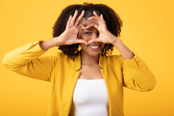 Красивая молодая черная леди, показывающая жест в форме сердца рядом с глазом и улыбающаяся, милая женщина, флиртующая на жёлтом фоне студии. Концепция жестов, эмоций - Фото, изображение