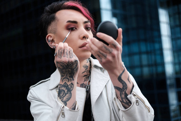 Androgynous male doing himself make outdoors. He is wearing a white leather jacket, pink hair and tattoos. Alternative lifestyle, edgy and pride concept. - Photo, Image