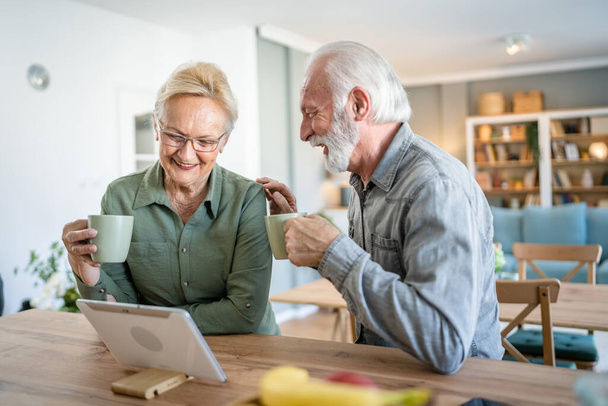 Senior couple caucasian old man and woman husband and wife pensioner grandfather and grandmother use digital tablet have cup of coffee at home in bright room real people family concept daily routine - Photo, Image