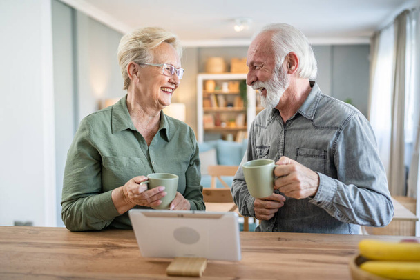 Senior couple caucasian old man and woman husband and wife pensioner grandfather and grandmother use digital tablet have cup of coffee at home in bright room real people family concept daily routine - Foto, afbeelding