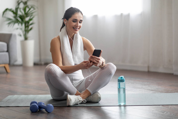 Music for fitness, mobile app. Cheerful pretty young athletic woman sitting on yoga mat, using smartphone and earphones, listening to music while exercising at home, copy space - Foto, Imagem